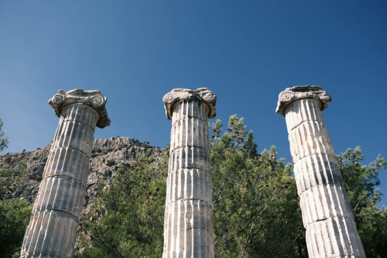 Three weathered stone columns from an ancient ruin, symbolizing the three levels of the Swiss tax system (federal, cantonal, communal), against a clear blue sky with a rocky hill and green trees in the background.